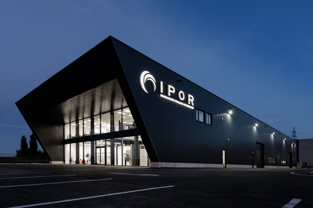 Moderne Firmenzentrale von IPOR bei Abenddämmerung. Das markante Gebäude mit futuristischer Architektur und großer Glasfront ist hell beleuchtet. Die leuchtende Beschriftung ‚IPOR – Innovativer Poolbau Ratzesberger‘ ist prominent an der Fassade angebracht. Der davorliegende Parkplatz ist leer.