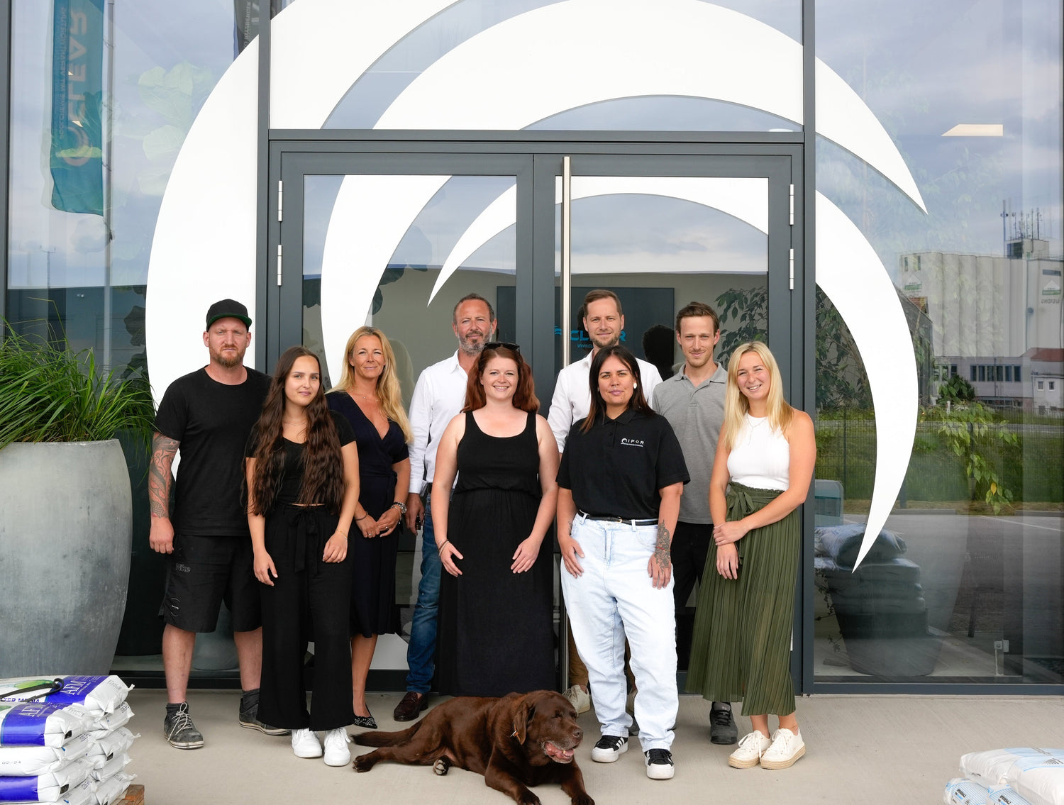 Gruppenfoto eines Teams vor einem modernen Firmengebäude mit großem, weißem Logo auf der Glasfassade. Zehn Personen, darunter Männer und Frauen in Freizeit- bis Businesskleidung, posieren gemeinsam mit einem braunen Labrador-Hund. Links und rechts sind Paletten mit Produkten gestapelt. Im Hintergrund sind Firmengebäude und Flaggen mit dem Schriftzug ‚CLEAR‘ sichtbar.