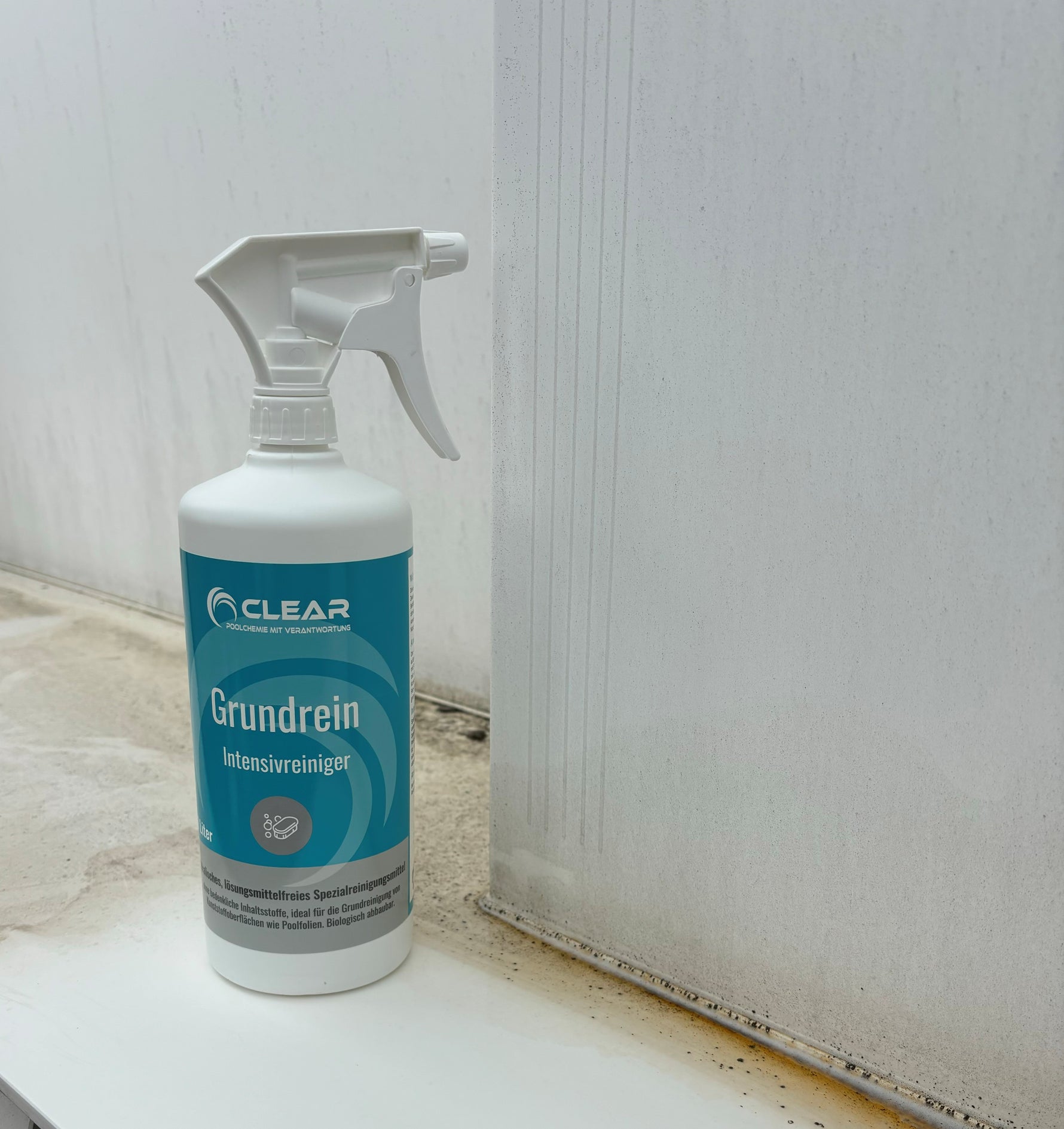 Flasche des CLEAR Grundrein Intensivreinigers mit Sprühkopf steht vor einer verschmutzten Poolwand. Die Wand zeigt deutlich sichtbare Ablagerungen und Reinigungsspuren, was die Wirksamkeit des Produkts demonstriert. Ideal zur Grundreinigung von Kunststoff- und Pooloberflächen.