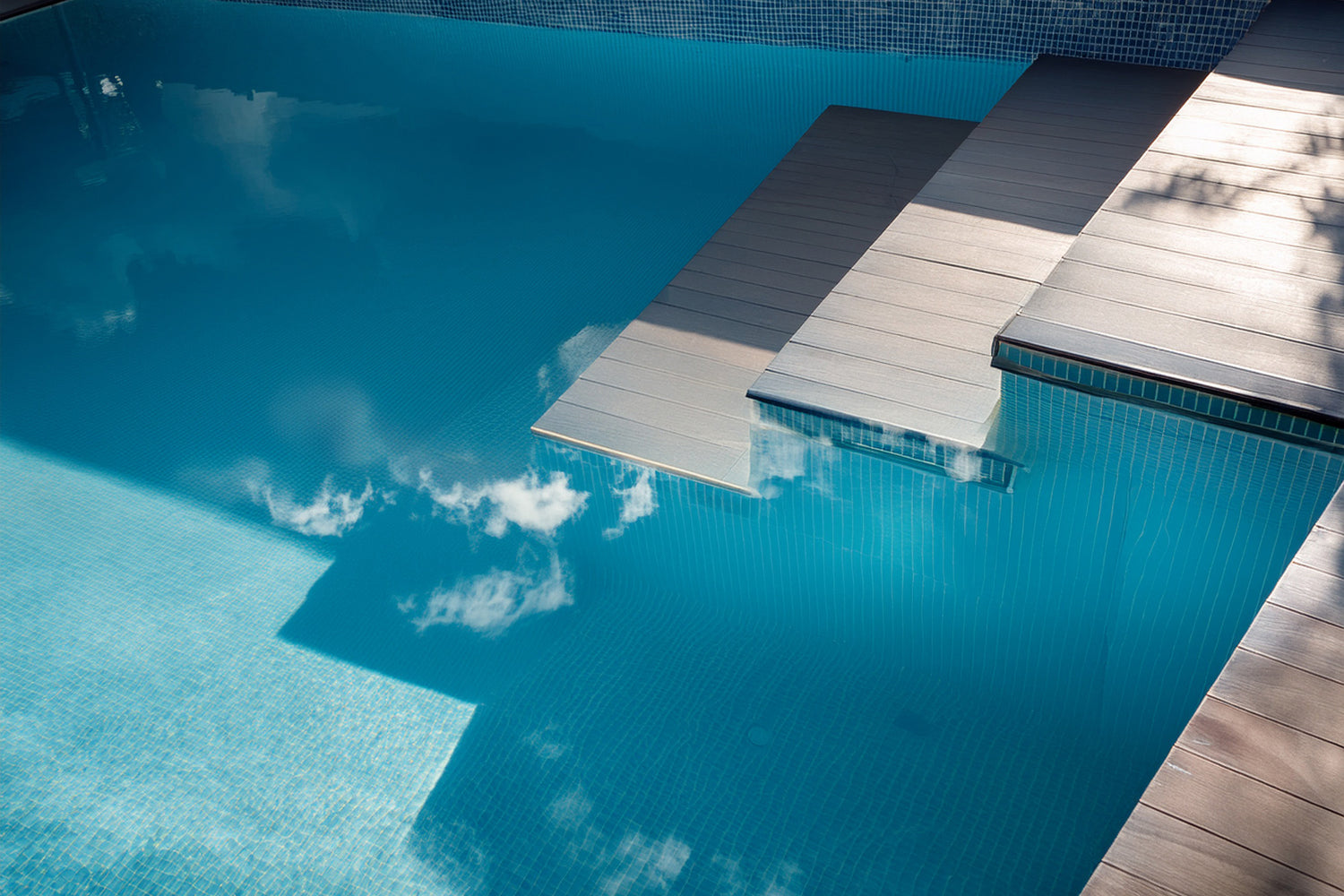 Detailaufnahme eines modernen Pools mit Mosaikfliesen und integrierten Stufen aus hellen Poolplatten. Auf der Wasseroberfläche spiegeln sich Wolken und Sonnenlicht, wodurch eine harmonische und hochwertige Ästhetik entsteht.